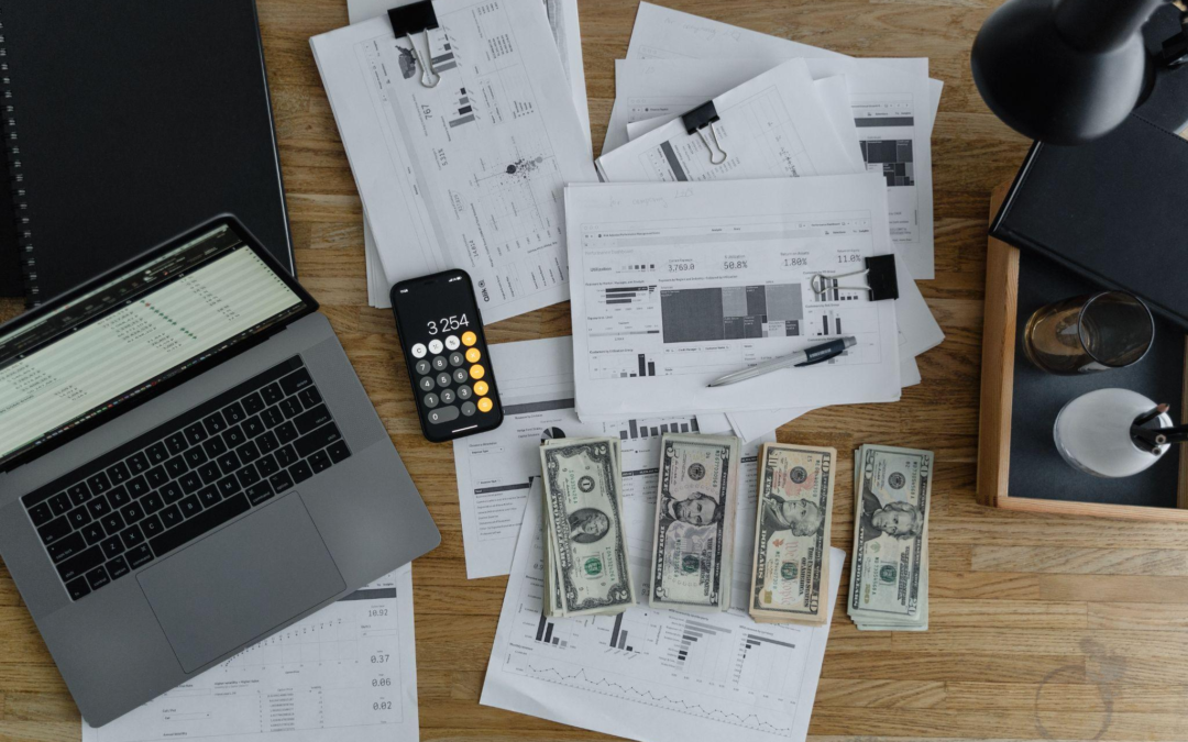 A laptop, some papers, dollars and a phone kept on a wooden table.