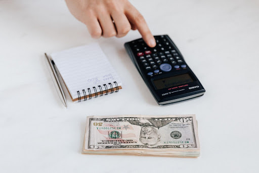 A hand putting some information into a calculator with a stack of American bills nearby.